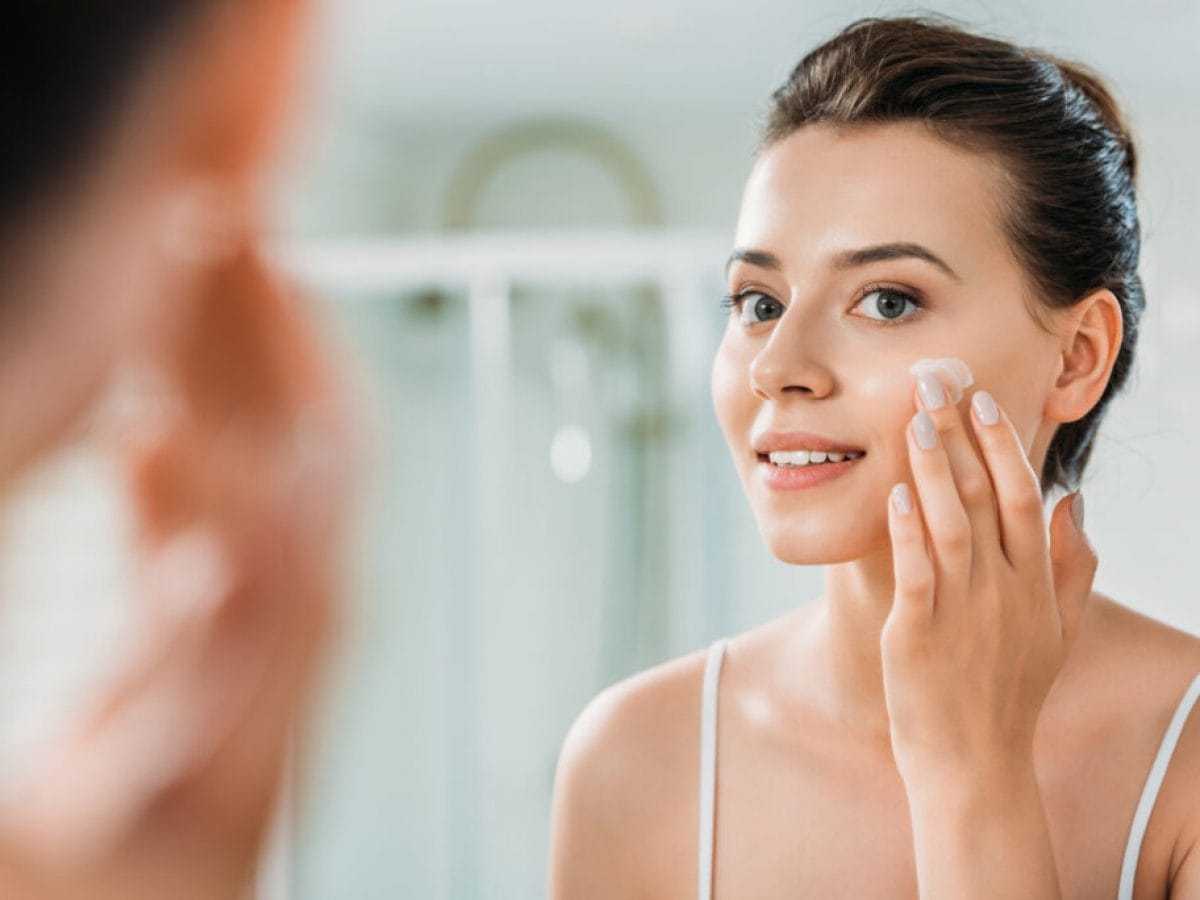 Woman applying moisturizer to her face in front of a bathroom mirror.