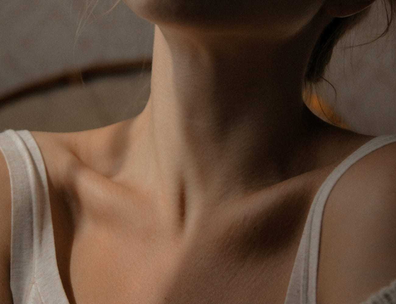 Close-up of a woman's neck and collarbone in soft lighting, wearing a white tank top.