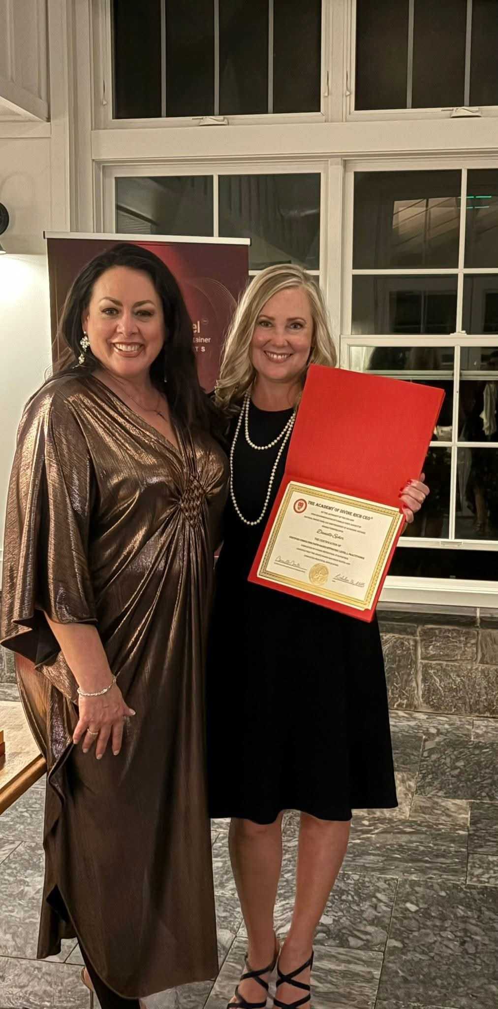 Two women smiling, one holding a red certificate in a formal setting.