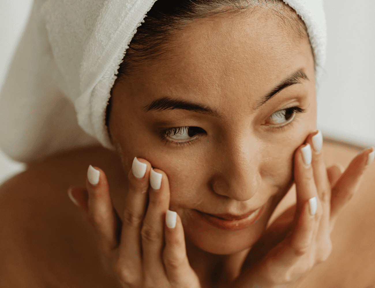 Woman with towel on head applying skincare to face.